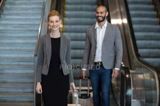 Улыбающиеся деловые люди с багажом спускаются на эскалаторе в терминале аэропорта — стоковое фото