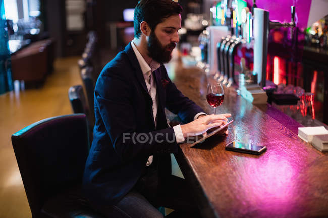 Empresário usando tablet digital com copo de vinho e telefone celular no balcão no bar — Fotografia de Stock
