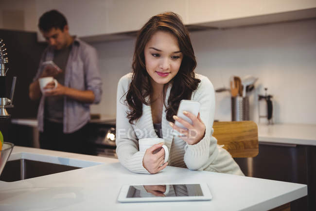 Woman using mobile phone while having coffee in kitchen at home — Stock Photo