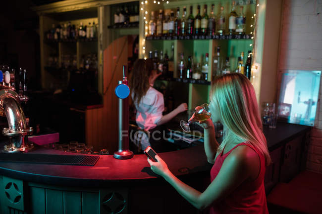 Donna che usa il telefono cellulare mentre beve un bicchiere di vino al bancone del bar — Foto stock