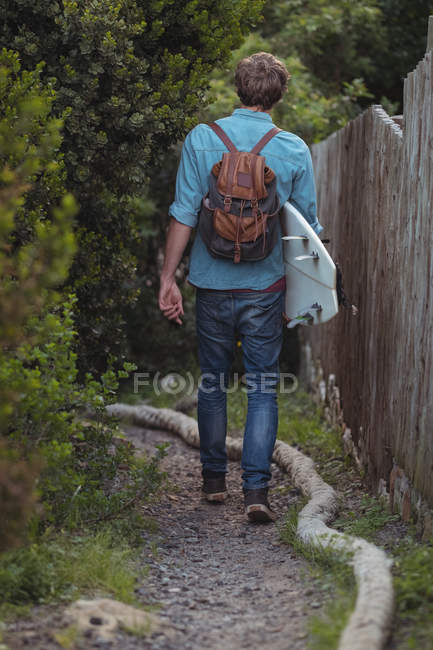 Visão traseira do homem com mochila carregando uma prancha de surf andando por uma trilha — Fotografia de Stock