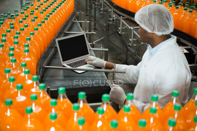 Vista de ángulo alto del trabajador masculino que utiliza el ordenador portátil en medio de la línea de producción en la fábrica de jugo - foto de stock