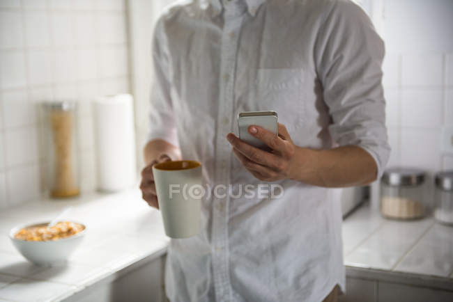 No meio da seção do homem usando telefone celular enquanto toma uma xícara de café em casa — Fotografia de Stock