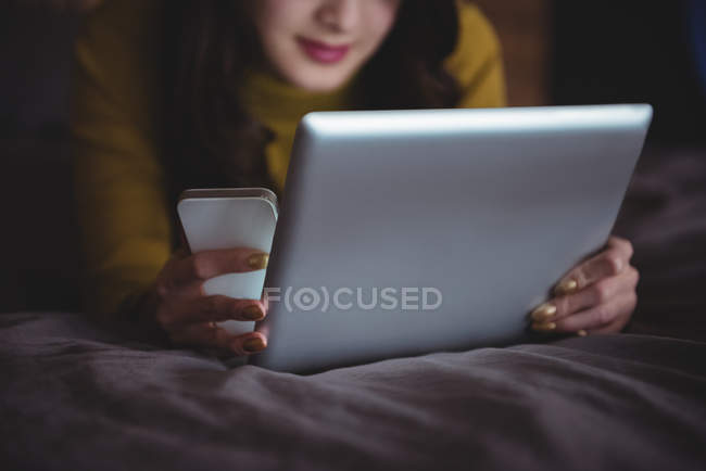 Жінка лежить на ліжку, використовуючи цифровий планшет і мобільний телефон в спальні вдома — стокове фото
