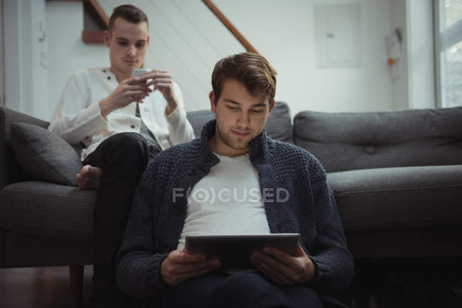 Gay couple using mobile phone and digital tablet in living room at home — Stock Photo