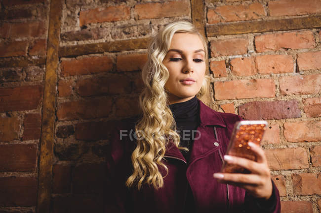 Schöne blonde Frau, die mit Handy gegen Ziegelmauer steht — Stockfoto