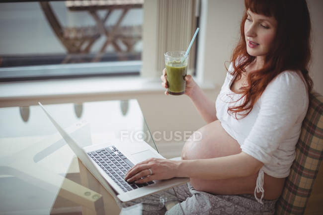 Mujer embarazada usando el ordenador portátil mientras tiene jugo en casa - foto de stock