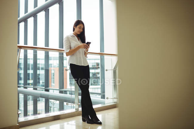 Бізнес-леді, спираючись на перил і використовуючи мобільний телефон в офісі — стокове фото