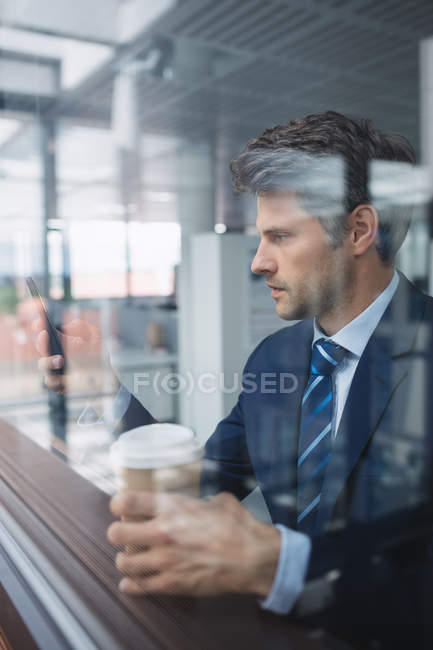 Empresário usando telefone celular e segurando copo de café descartável no escritório — Fotografia de Stock
