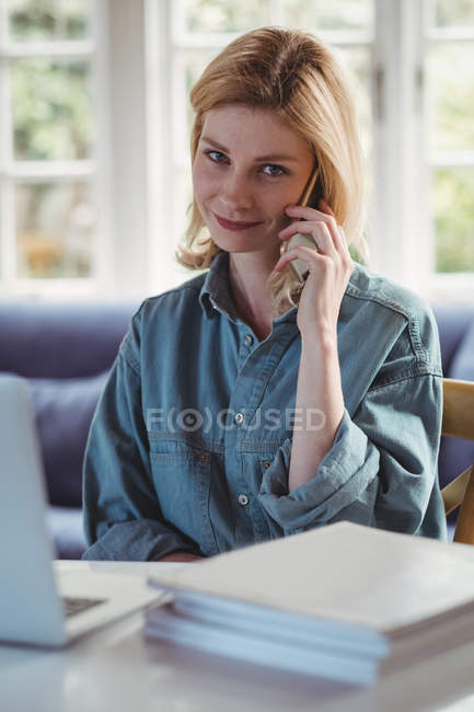 Красива жінка розмовляє на мобільному телефоні, використовуючи ноутбук у вітальні вдома — стокове фото