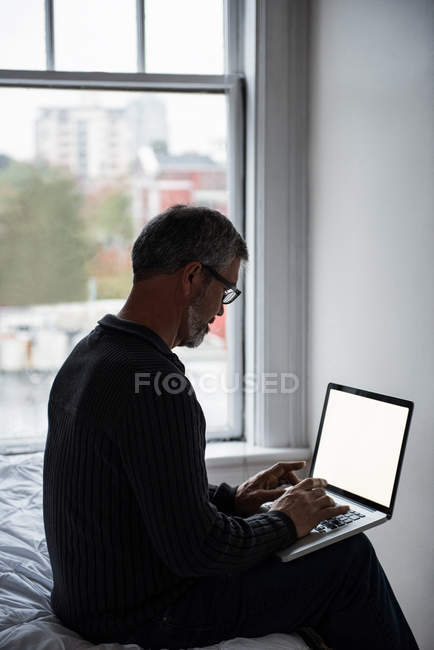 Чоловік сидить на ліжку і використовує ноутбук вдома — стокове фото
