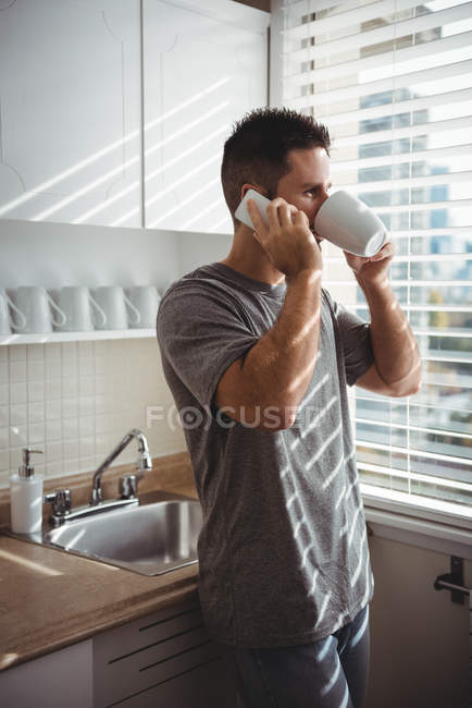 Hombre hablando por teléfono móvil mientras toma café en la cocina en casa - foto de stock