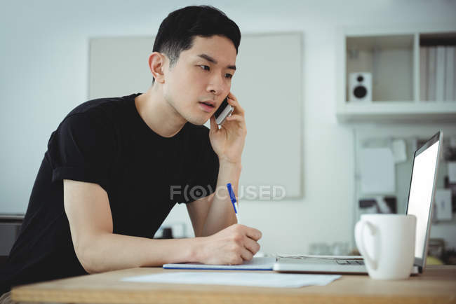 Business executive writing on diary while talking on mobile phone in office — Stock Photo