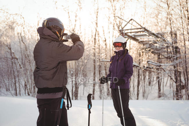 Homem de esqui fotografando mulher com telefone celular em estância de esqui — Fotografia de Stock