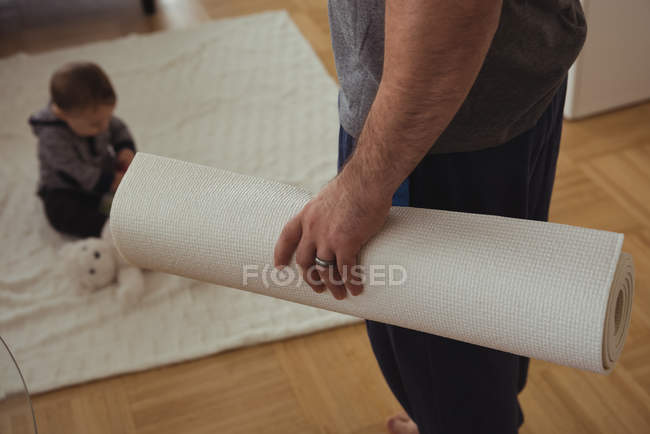 Pai segurando tapete de exercício enquanto bebê brincando no fundo em casa — Fotografia de Stock