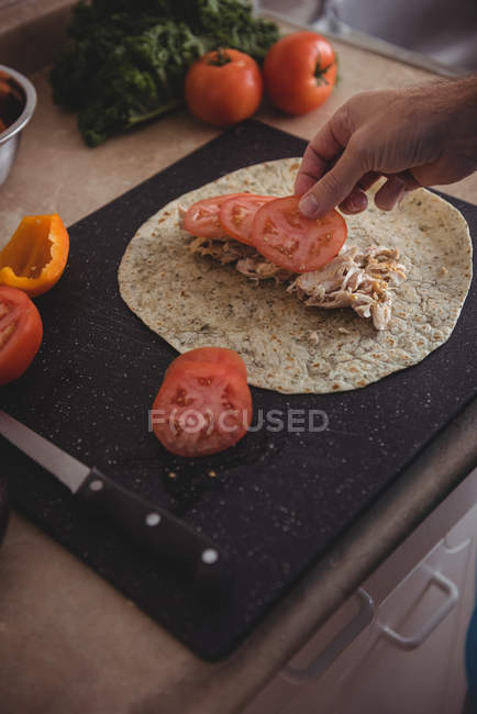 Primer plano de la mano masculina colocando rodajas de tomate en el burrito en la cocina en casa - foto de stock