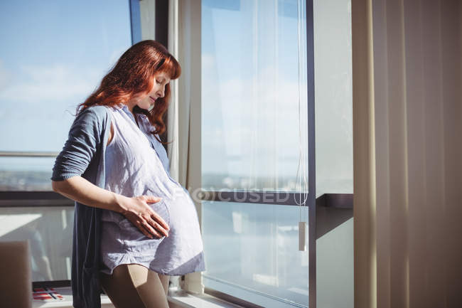 Задумчивая беременная женщина стоит у окна в гостиной дома — стоковое фото