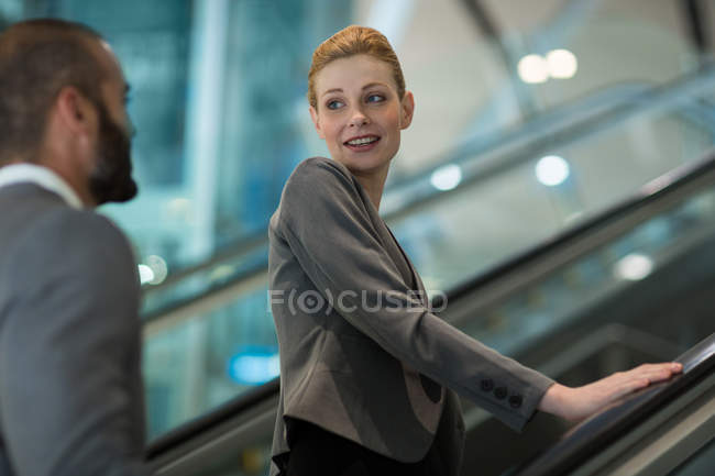 Gente de negocios interactuando entre sí mientras suben en escaleras mecánicas en la terminal del aeropuerto - foto de stock