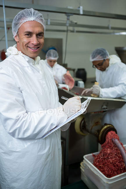 Butcher writing on clipboard while coworkers placing meat in mincing machine at meat factory — Stock Photo