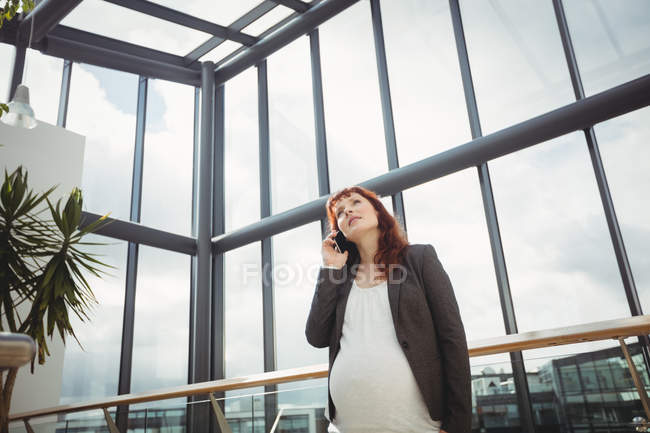 Беременная деловая женщина разговаривает по мобильному телефону возле коридора в офисе — стоковое фото