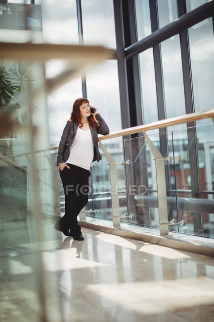Беременная деловая женщина, использующая мобильный телефон рядом с коридором в офисе — стоковое фото