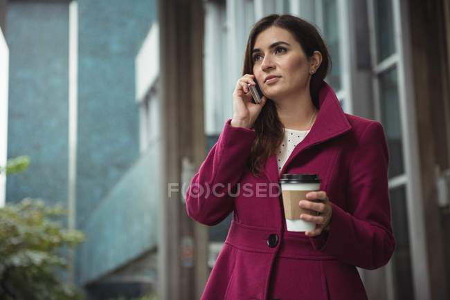 Бізнес-леді тримає одноразову чашку кави і розмовляє на мобільному телефоні біля офісної будівлі — стокове фото