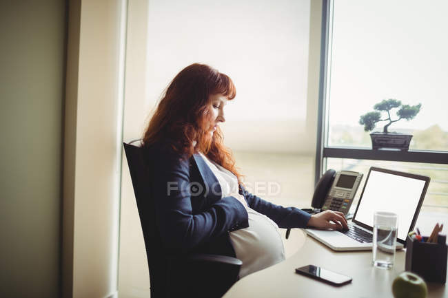 Вагітна бізнес-леді торкається живота, використовуючи ноутбук в офісі — стокове фото