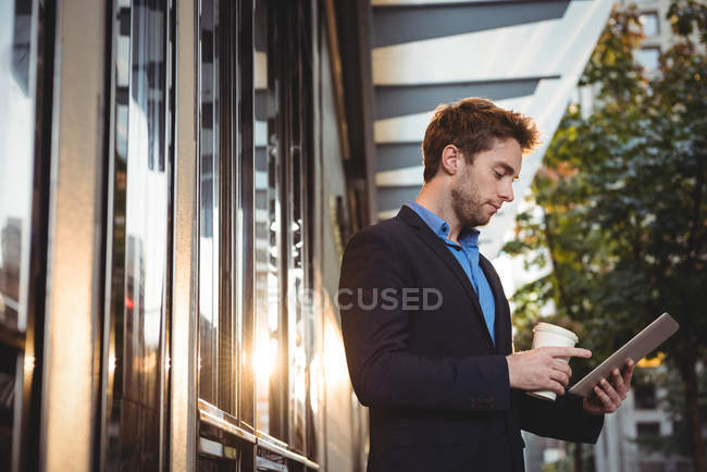 Бізнесмен тримає одноразову чашку кави і використовує цифровий планшет, стоячи на вулиці — стокове фото