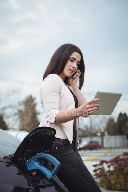 Красива жінка говорити на мобільному телефоні під час зарядки електричний автомобіль на вулиці — стокове фото