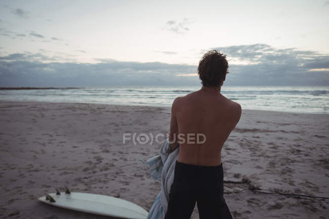 Человек с видом сзади, стоящий на пляже с доской для серфинга в сумерках — стоковое фото