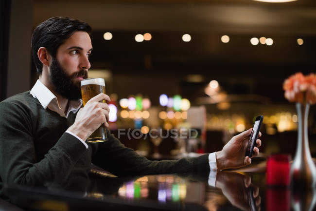 Hombre sosteniendo el teléfono móvil y tomando un vaso de cerveza en el bar - foto de stock