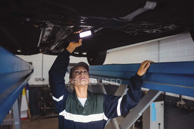 Mécanicien féminin examinant une voiture avec lampe de poche dans le garage de réparation — Photo de stock