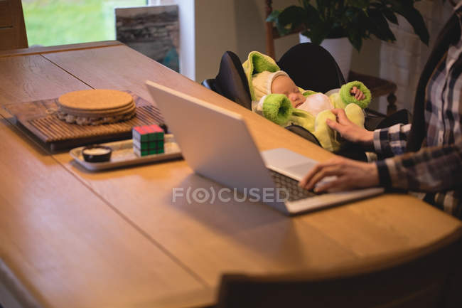 Sección media de la madre usando el ordenador portátil mientras cuida al bebé en casa - foto de stock
