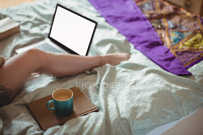 Женщина на кровати с ноутбуком и чашкой кофе дома — стоковое фото