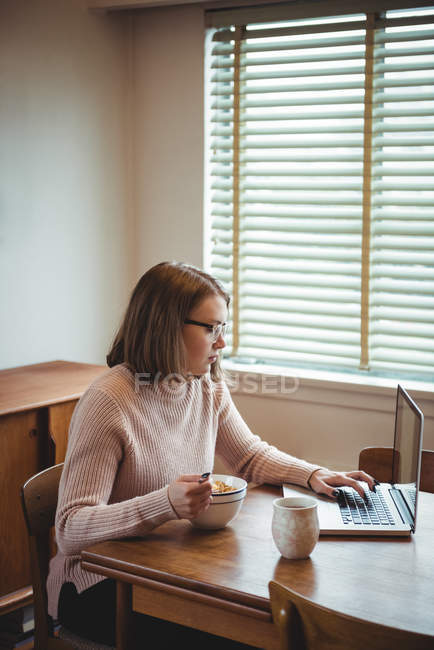 Femme utilisant un ordinateur portable tout en prenant le petit déjeuner dans le salon à la maison — Photo de stock