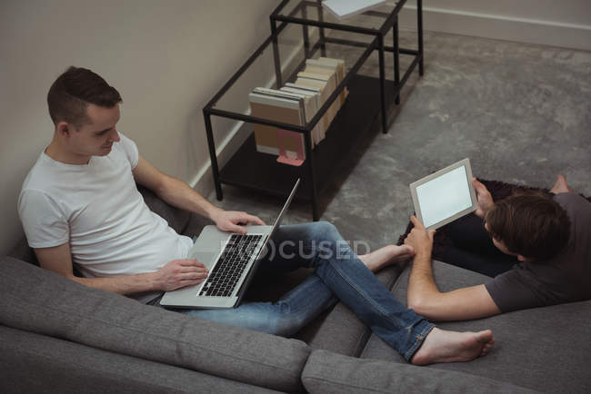 Гей пара за допомогою цифрових планшетів і ноутбук у вітальні на дому — стокове фото