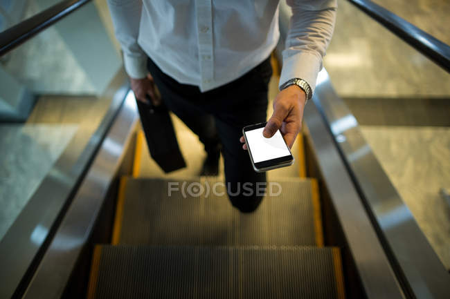 Sección media del hombre que utiliza el teléfono móvil en la escalera mecánica en el aeropuerto - foto de stock