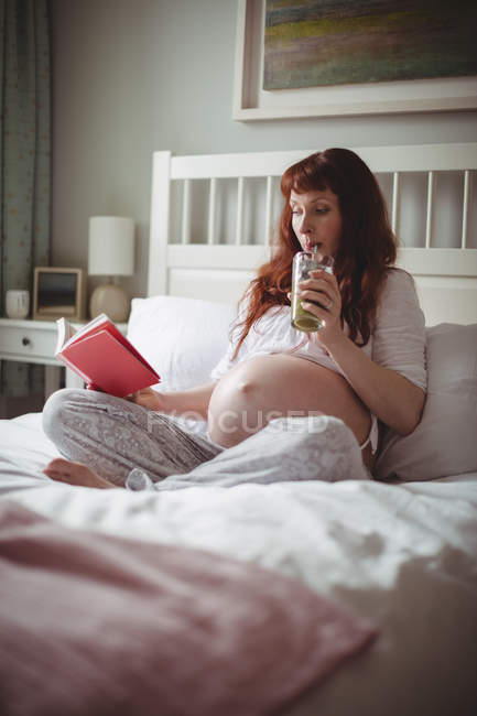 Donna incinta che beve succo mentre legge libro sul letto in camera da letto — Foto stock