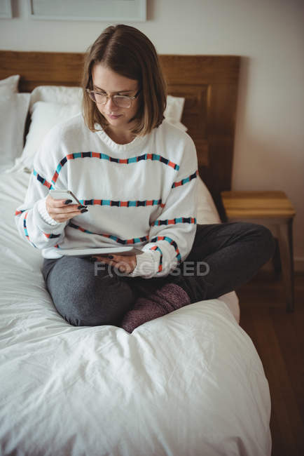 Mulher sentada na cama usando celular e tablet digital no quarto — Fotografia de Stock