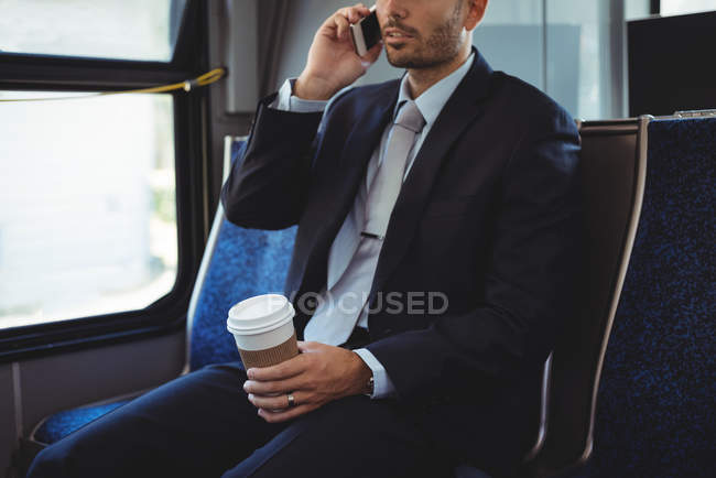 Empresário segurando uma xícara de café descartável falando no telefone celular enquanto viaja em ônibus — Fotografia de Stock