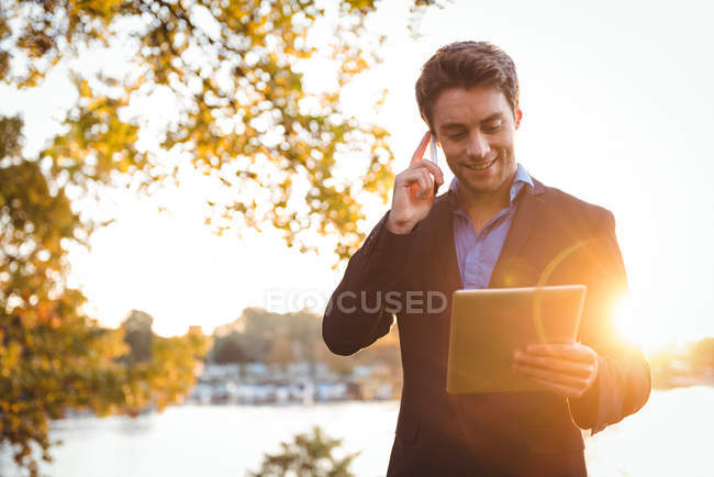 Бизнесмен разговаривает по мобильному телефону во время использования цифрового планшета в парке на солнце — стоковое фото