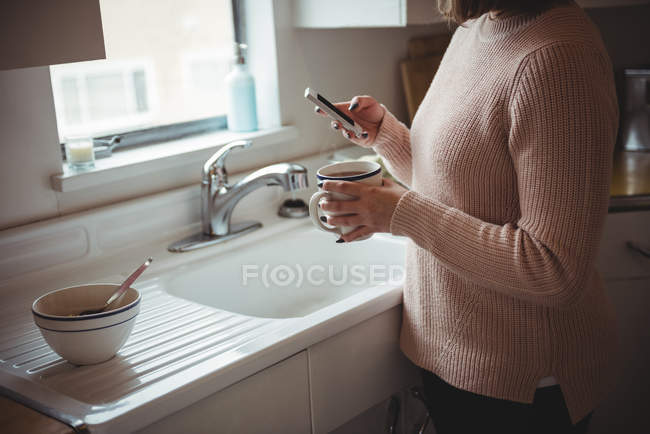 Mujer usando el teléfono móvil mientras sostiene la taza de café en la cocina en casa - foto de stock