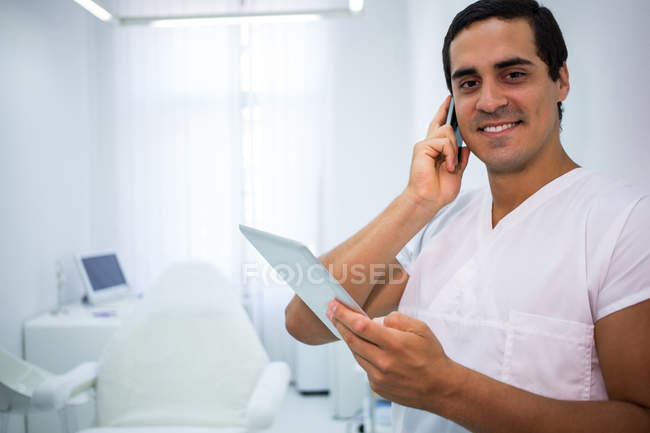 Dentista falando no celular e segurando tablet digital na clínica — Fotografia de Stock