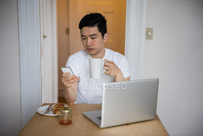 Uomo che utilizza il telefono cellulare mentre prende una tazza di caffè a casa — Foto stock