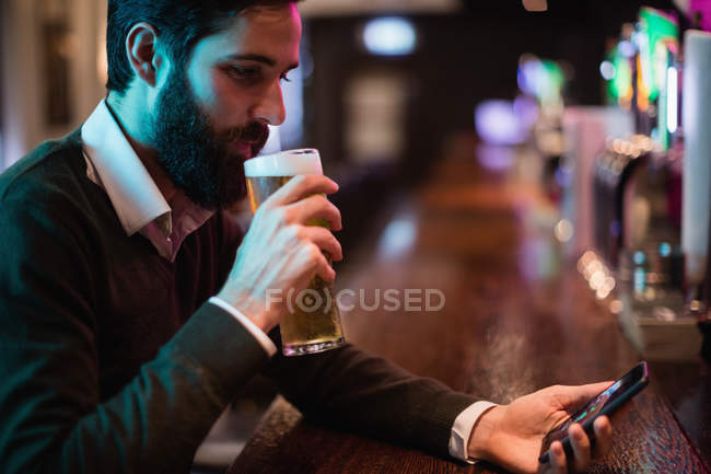 Homem olhando para o telefone celular enquanto toma um copo de cerveja no balcão do bar — Fotografia de Stock