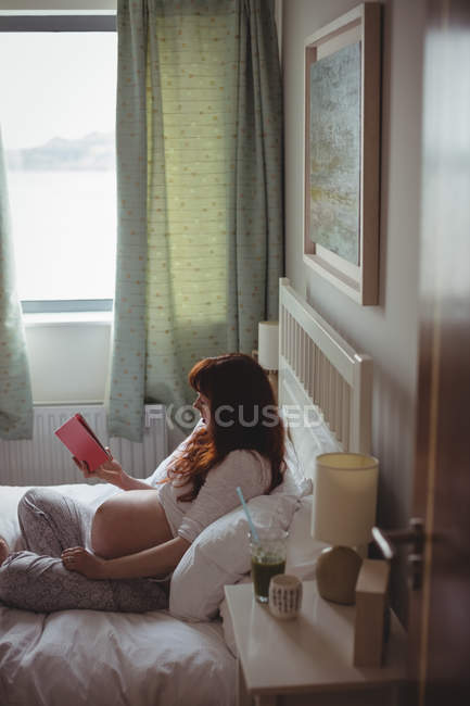 Pregnant woman reading book on bed in bedroom — Stock Photo