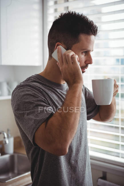 Чоловік розмовляє на мобільному телефоні, маючи каву на кухні вдома — стокове фото