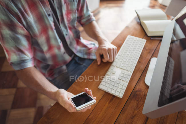 Partie médiane d'un homme assis au bureau de l'ordinateur et regardant le téléphone mobile — Photo de stock