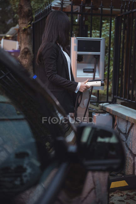 Partie médiane de la femme tenant le chargeur de voiture à la station de charge du véhicule électrique — Photo de stock