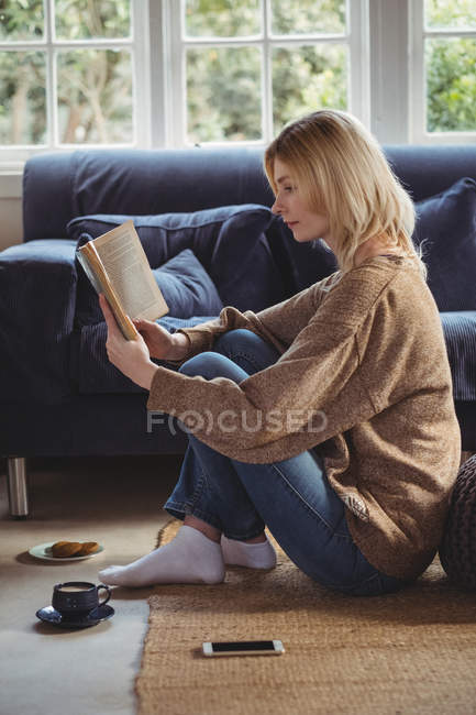 Beautiful woman reading book while having tea in living room at home — Stock Photo